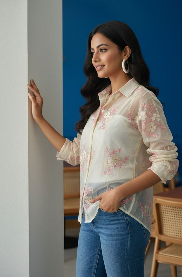Woman wearing a white floral blouse and blue jeans standing indoors against a blue wall.